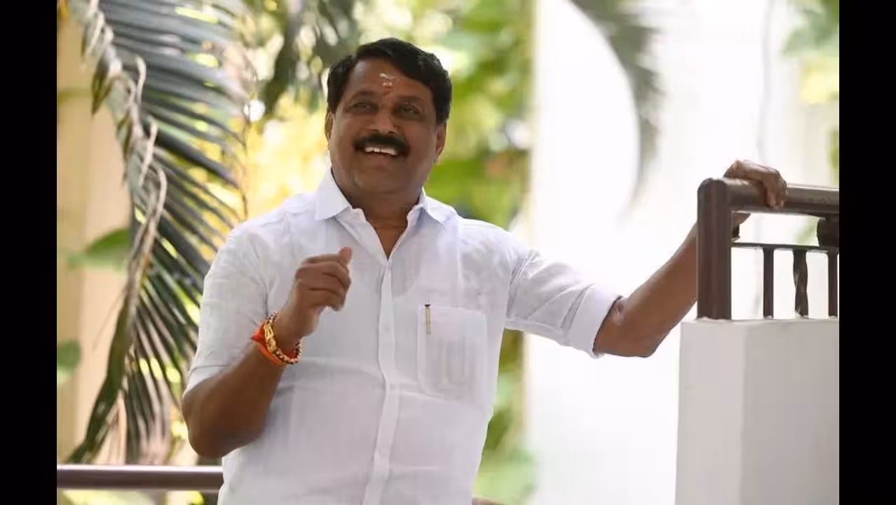 Nainar Nagendran laughing while standing on a balcony surrounded by greenery, in a white shirt
