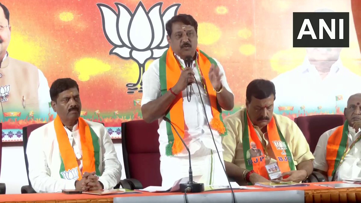 Nainar Nagendran standing and addressing a party meeting from behind a table, with colleagues seated on either side and a lotus backdrop behind
