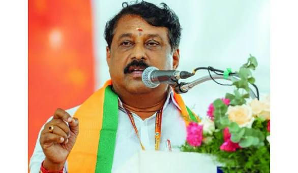 Nainar Nagendran speaking from a podium microphone, gesturing with one hand, wearing a party shawl
