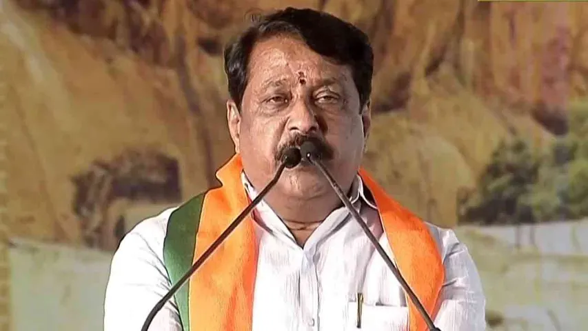 Nainar Nagendran addressing a gathering from a microphone stand, wearing a party shawl, with a large painted backdrop behind him