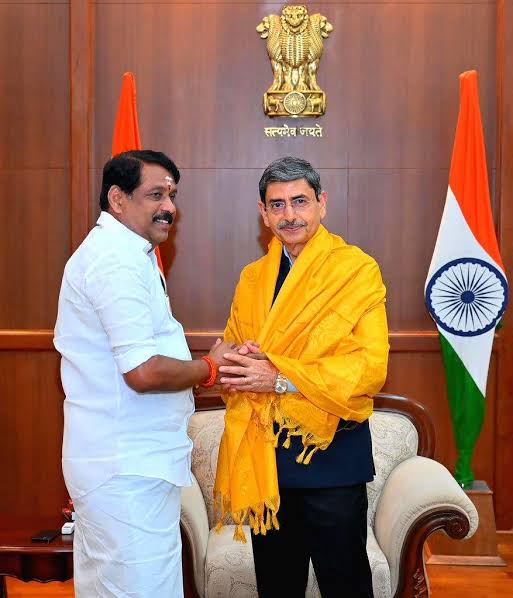 Nainar Nagendran shaking hands with the Governor of Tamil Nadu, who wears a yellow ceremonial shawl, in an office with the national emblem and flag behind them